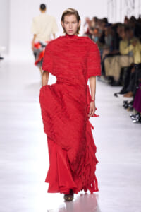 Model on a runway wearing a bright red textured gown with layered fringe and an oversized cape-like top.