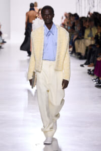 Male model walks a fashion runway in a cream boucle jacket, light blue shirt, and oversized cream trousers, clutch in hand.
