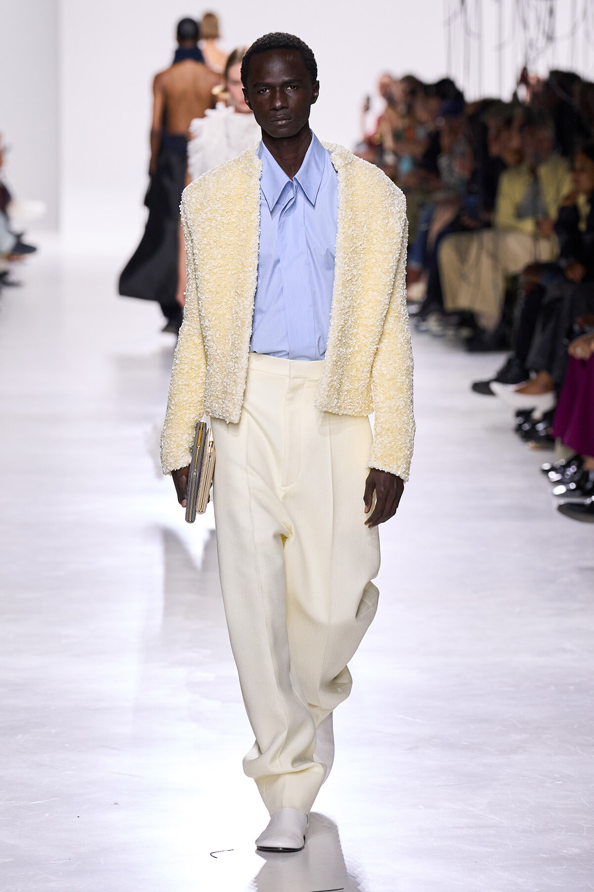 Male model walks a fashion runway in a cream boucle jacket, light blue shirt, and oversized cream trousers, clutch in hand.