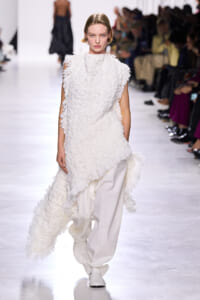 Model walking the runway in a white textured, fringed dress with loose white trousers, against a seated audience backdrop.