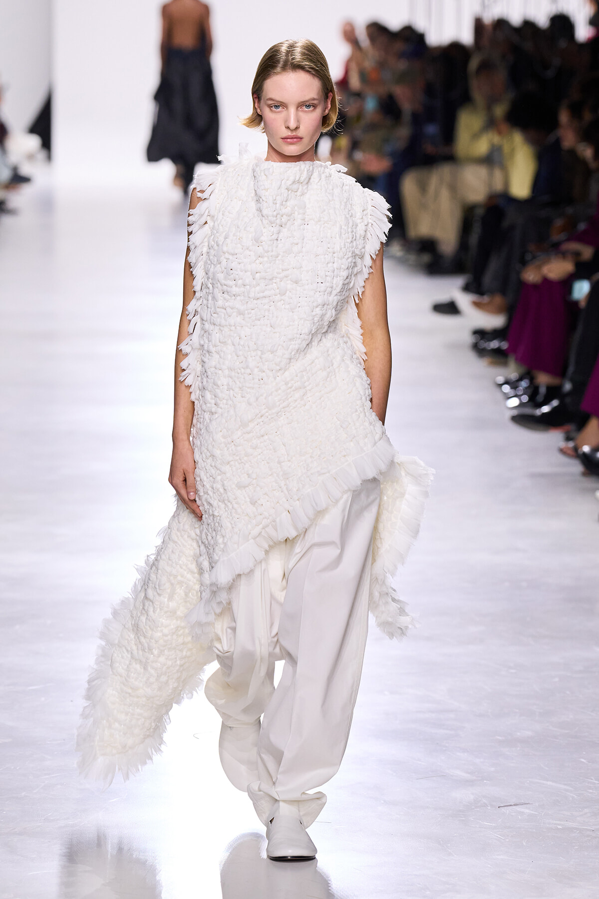 Model walking the runway in a white textured, fringed dress with loose white trousers, against a seated audience backdrop.