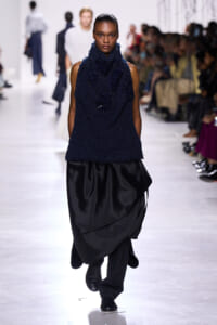 Model walks the runway in a navy textured sleeveless top with a large, draped black skirt; audience seated along the catwalk.