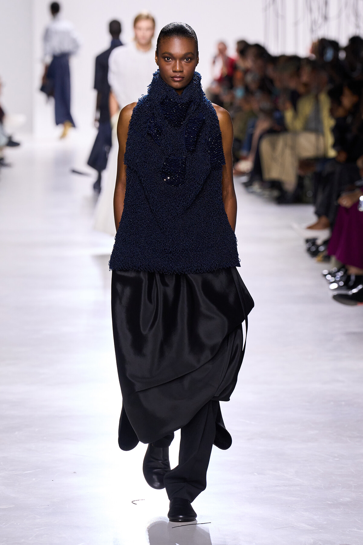 Model walks the runway in a navy textured sleeveless top with a large, draped black skirt; audience seated along the catwalk.