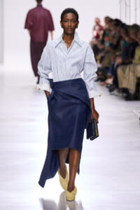 Model walking the runway in a light blue striped shirt with oversized sleeves and a dark blue wrap skirt, wearing yellow shoes and carrying a clutch