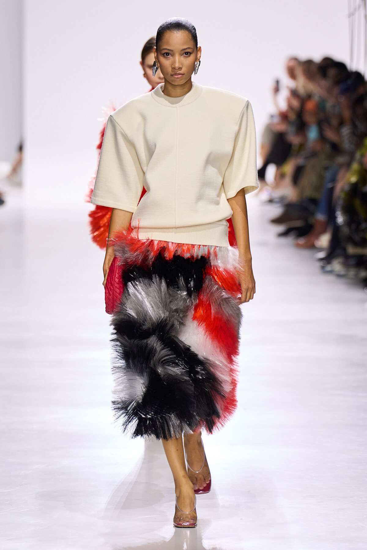 Model walks the runway in a cream oversized top and a knee‑length multicolored fur skirt (red, black, white) with clear heels.