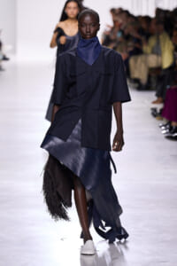 Model walks the runway in a navy oversized shirt dress with a blue neck scarf and a layered, fringed navy skirt/bag combo. White shoes visible.
