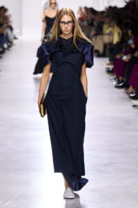 Model walking a runway in a navy long dress with dramatic satin puff sleeves, black lace yoke, and white shoes, holding a gold clutch.