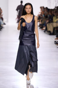 Model walking a fashion show in a navy sleeveless leather dress with a layered ruffled hem, carrying a fringed black bag and wearing white shoes.