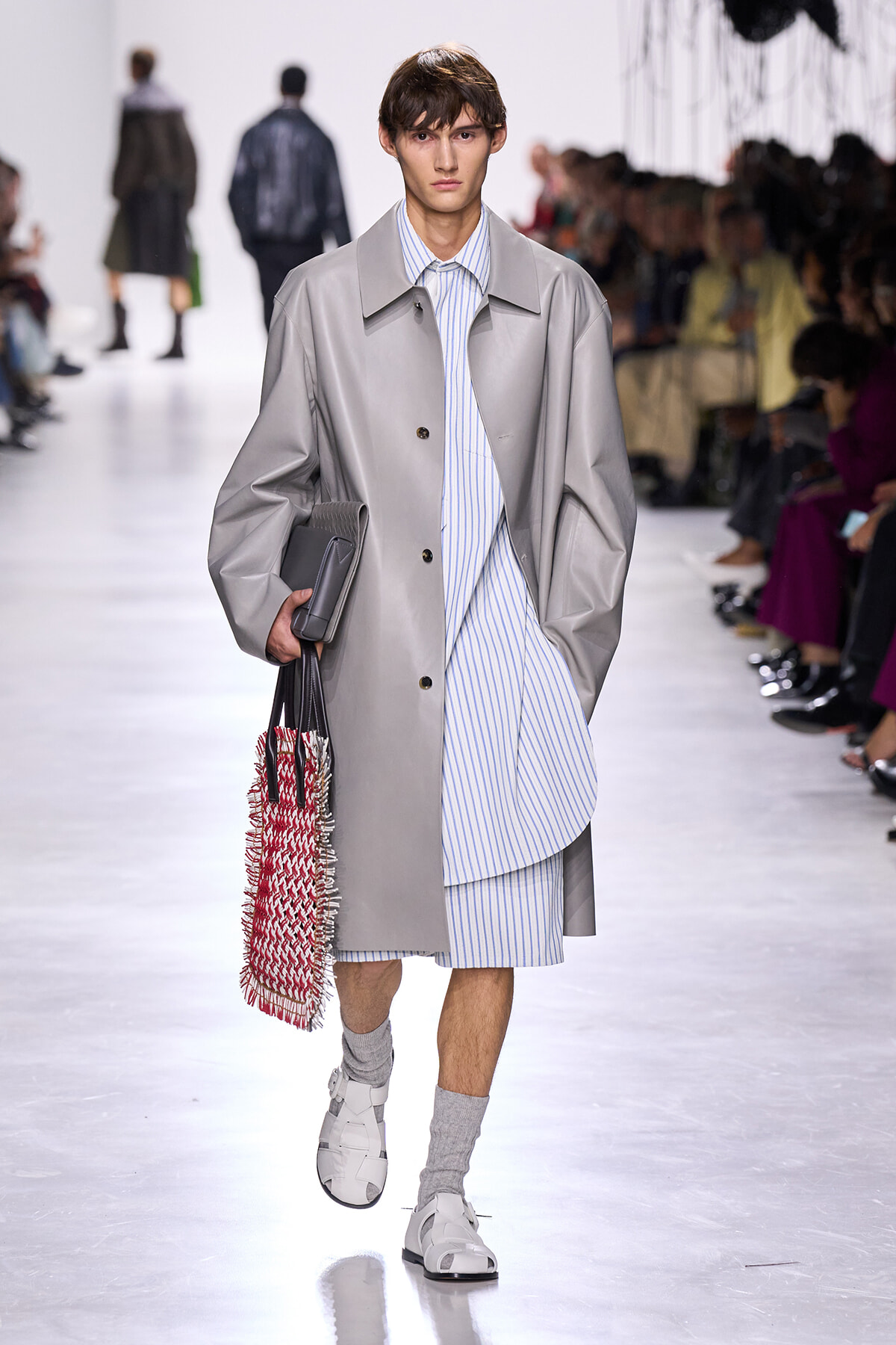 Male fashion model walks a runway in a light gray oversized coat over a blue-striped shirt and shorts, carrying a red-and-white woven bag and a gray clutch.