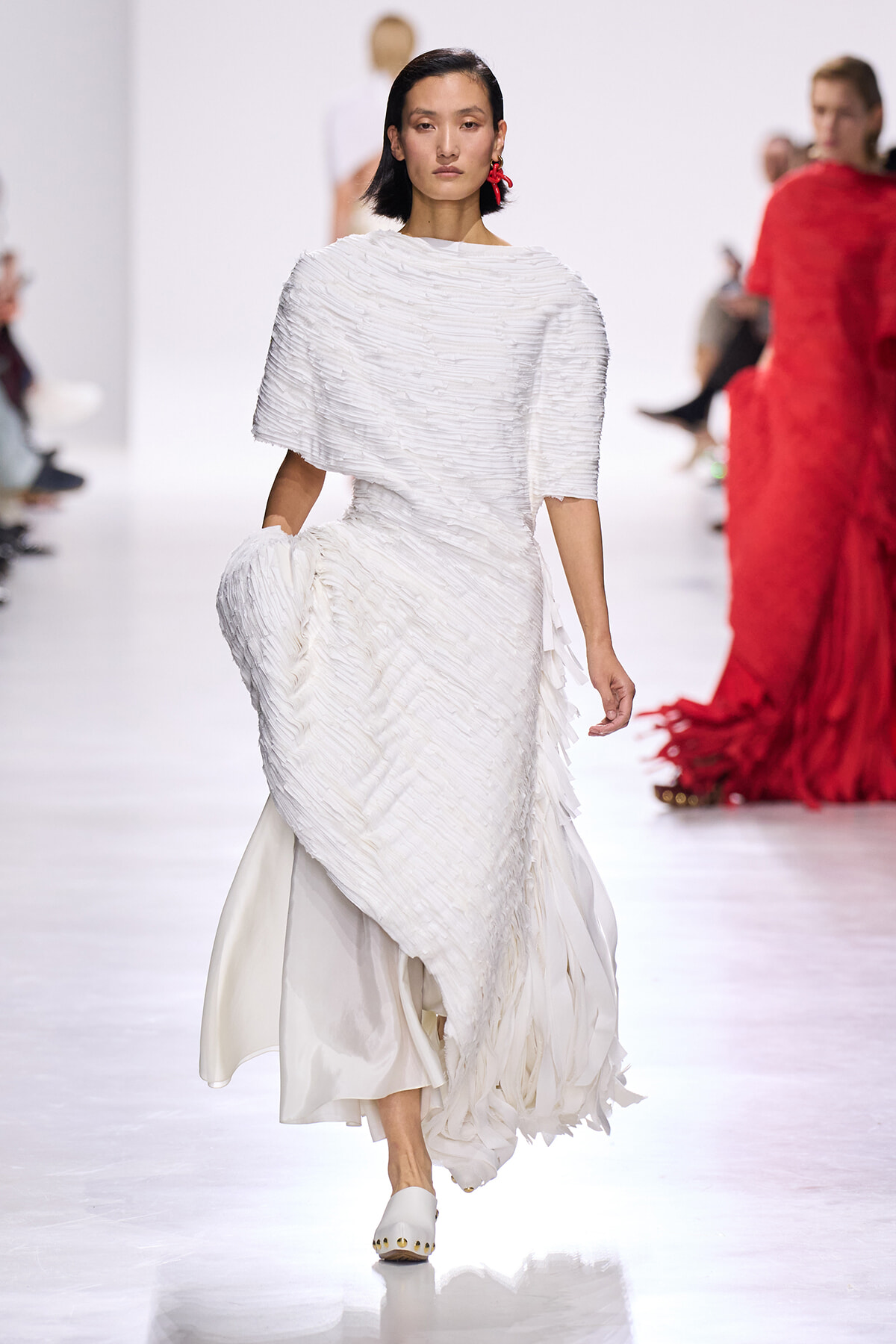 Model walking a fashion runway in a long white textured dress with fringe, paired with white shoes and red earring accents.