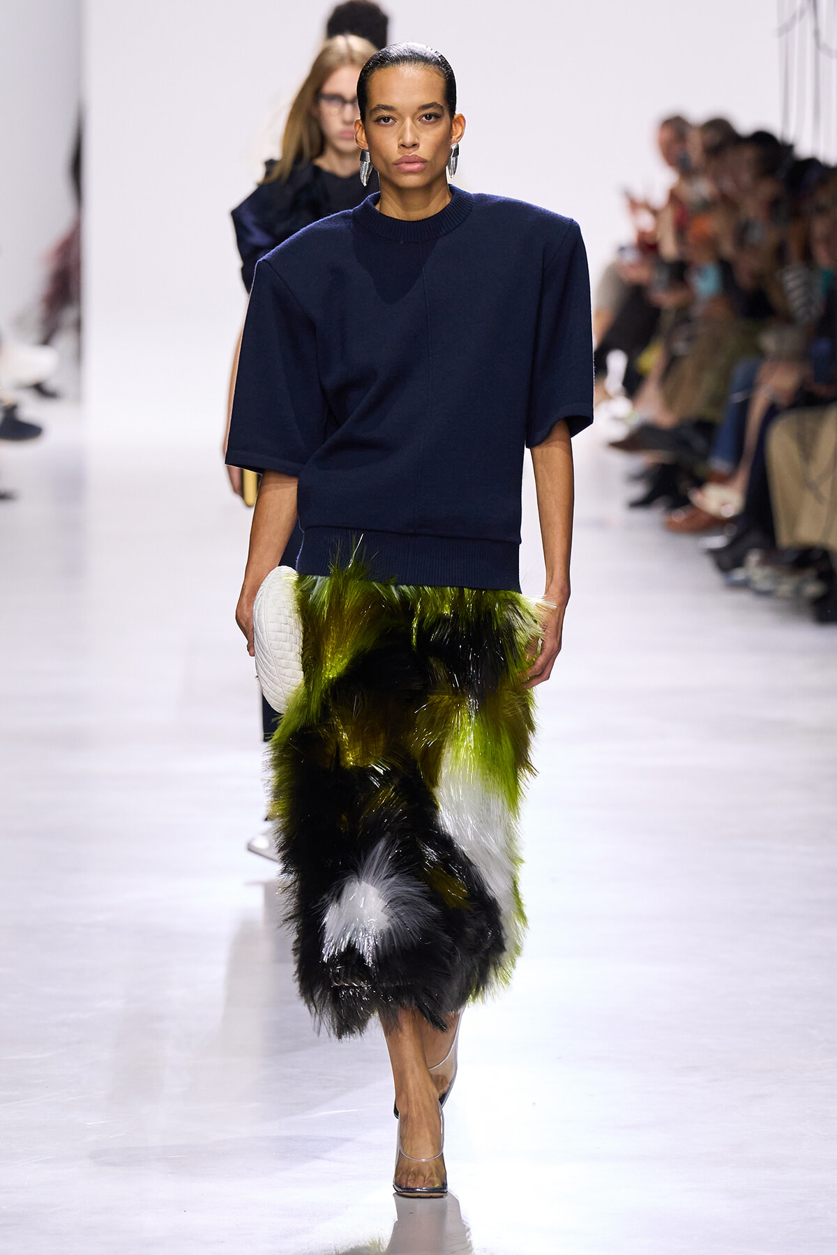Model walks runway in navy oversized top and green‑black fur skirt, carrying a white quilted clutch.