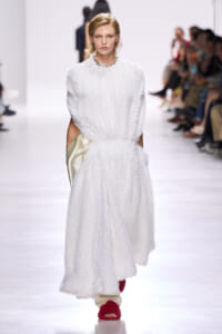 Model walks the runway in a white textured maxi dress with pockets and a beaded necklace, wearing red fuzzy slides.