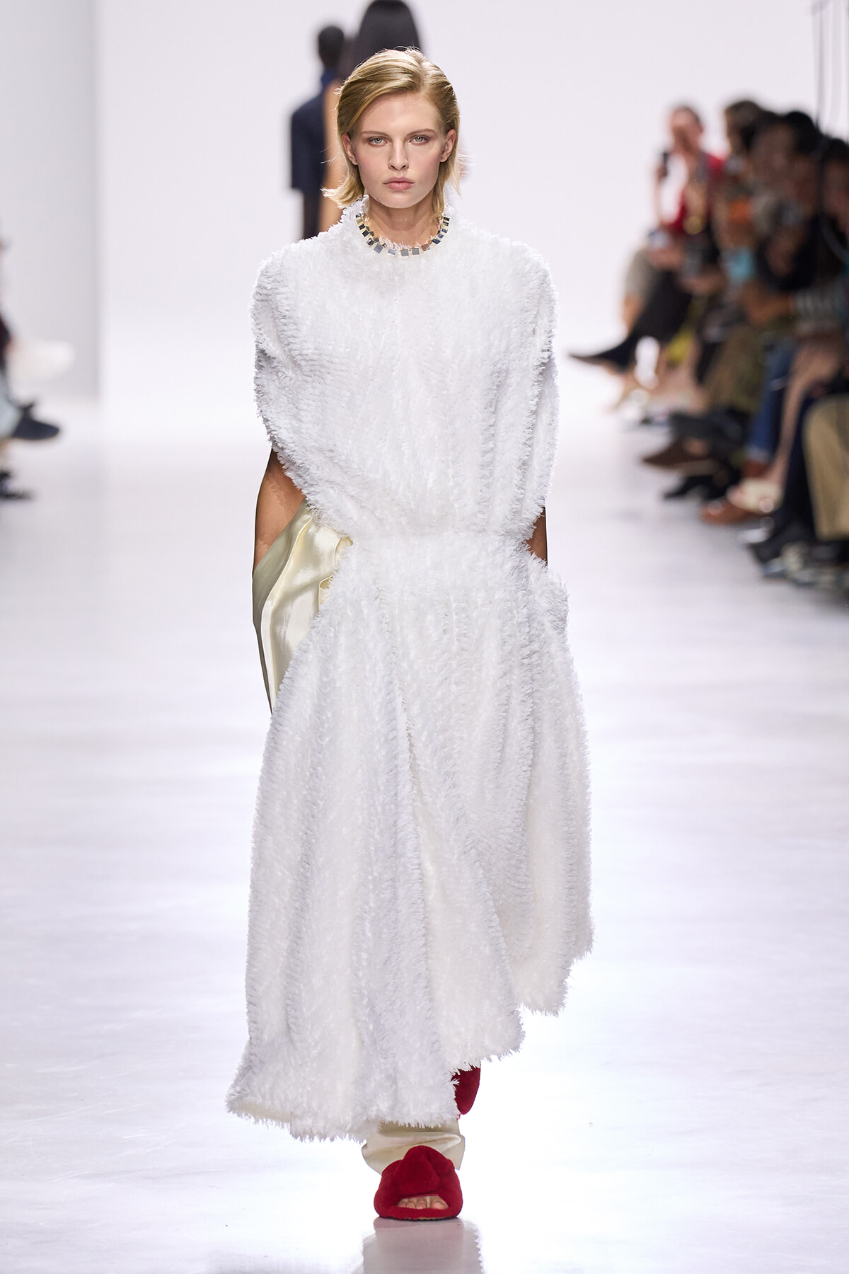 Model walks the runway in a white textured maxi dress with pockets and a beaded necklace, wearing red fuzzy slides.