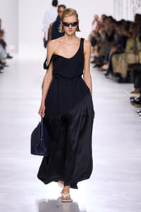 Model walks the runway in a black, one-shoulder dress, wearing sunglasses and carrying a blue textured handbag.”,