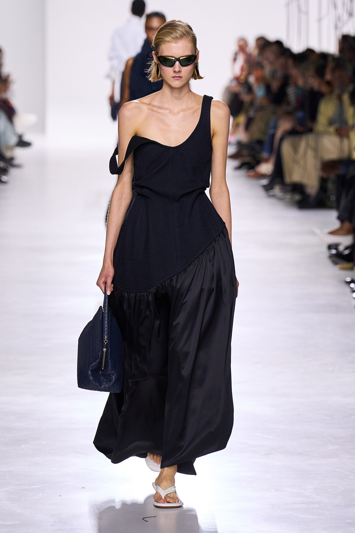 Model walks the runway in a black, one-shoulder dress, wearing sunglasses and carrying a blue textured handbag.”,