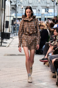 Model in brown plaid matching top and shorts with a large ruffled collar walks a fashion runway lined with seated spectators outside a venue.