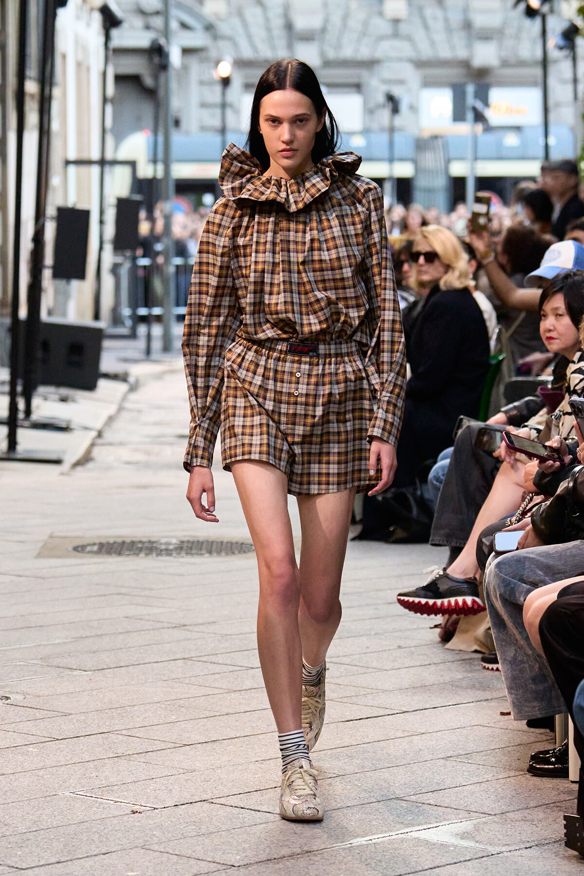 Model in brown plaid matching top and shorts with a large ruffled collar walks a fashion runway lined with seated spectators outside a venue.