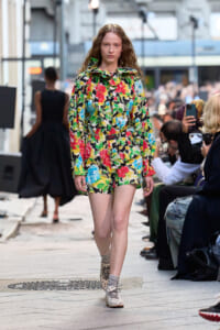 Female model walks on a city street runway wearing a bold multicolor floral blouse and matching shorts.
