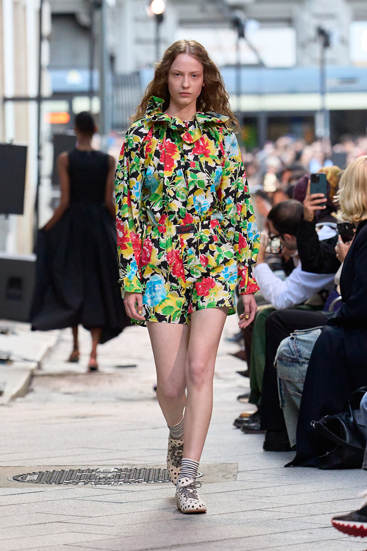 Female model walks on a city street runway wearing a bold multicolor floral blouse and matching shorts.