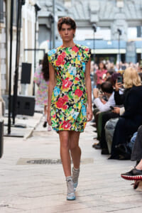 Model striding down a city street runway in a vivid floral shift dress with striped socks and pale blue shoes, spectators lining the sidewalk.