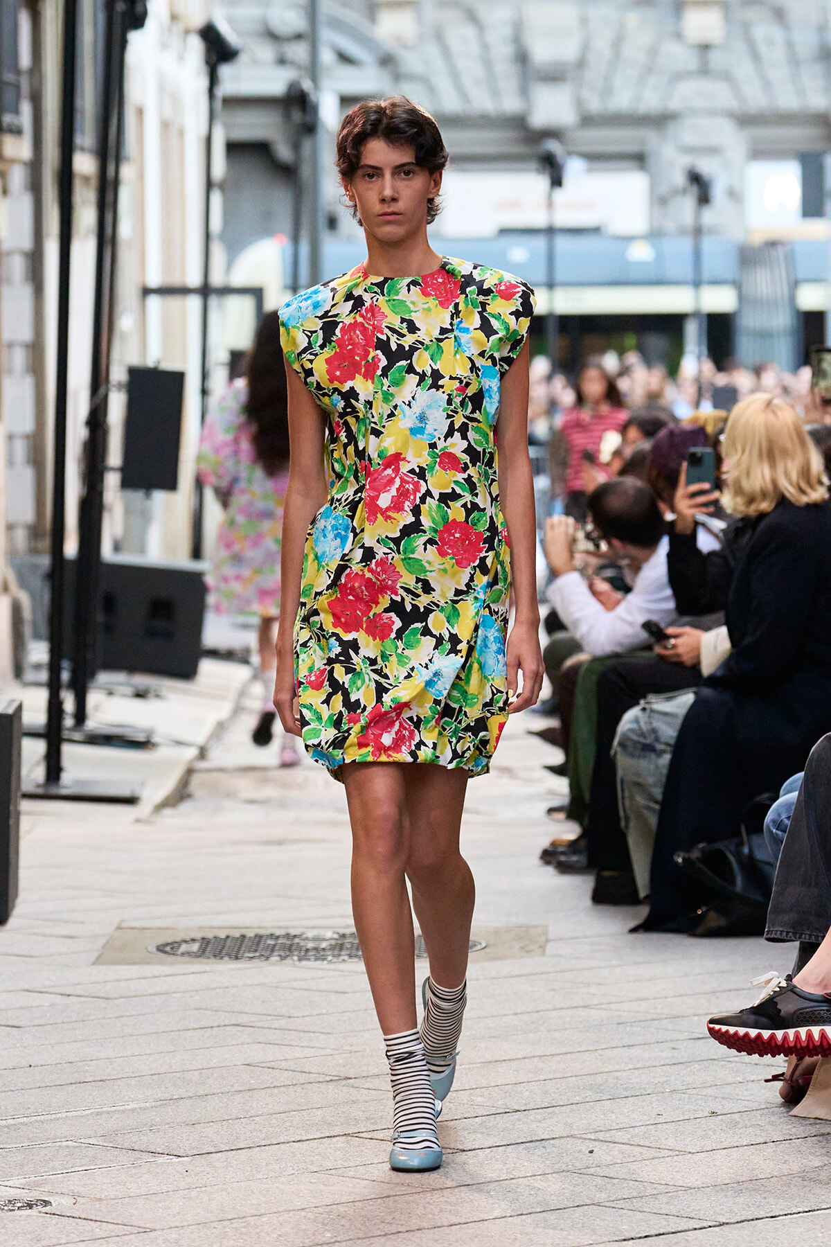 Model striding down a city street runway in a vivid floral shift dress with striped socks and pale blue shoes, spectators lining the sidewalk.