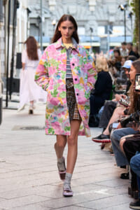 Model walking a street-style runway in a pink floral coat over a striped top and plaid skirt, with seated spectators lining the street.