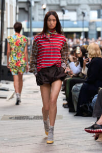 Model walks an urban runway wearing a red striped polo, brown puff shorts, and striped socks amid seated spectators and photographers.
