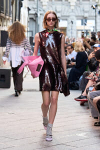 Model walking a street-style runway in a burgundy sequin shift dress with a pink oversized clutch and striped socks, outdoors.