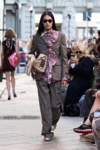 Model walking a city-style runway in a gray plaid suit with pink snake-print ruffles and matching scarf, wearing dark sunglasses.
