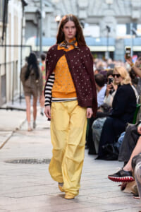 Female model walks a city-style runway wearing yellow wide pants, mustard top with studs, striped long sleeve underneath, maroon studded cardigan, and orange scarf.