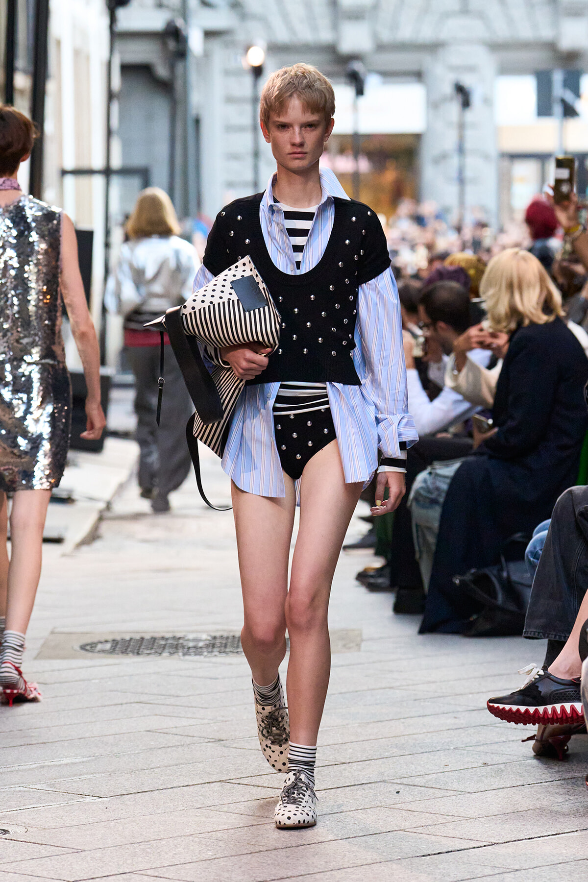 Model walking a city runway in a black studded vest over a blue striped shirt with black polka-dot bottoms, holding a striped clutch.