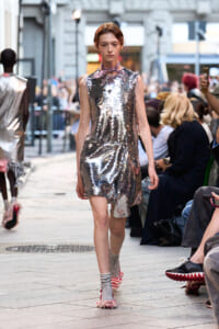 Model walking a city-fashion runway in a silver sequin sheath, pink neck scarf and striped socks, spectators on both sides.