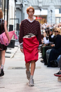 Model walking a street-fashion runway in a burgundy studded sweater, white collar shirt, red satin skirt, striped socks, and zebra-print shoes in front of spectators.