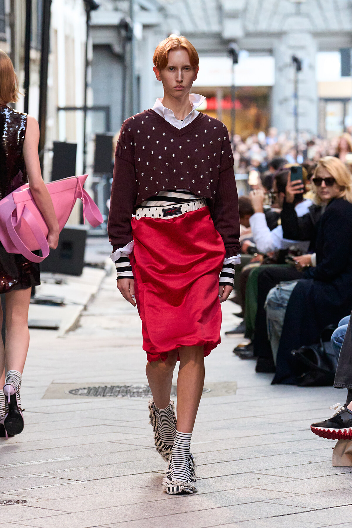 Model walking a street-fashion runway in a burgundy studded sweater, white collar shirt, red satin skirt, striped socks, and zebra-print shoes in front of spectators.