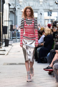 Model walking a street runway in a bold red-and-white striped outfit with a metallic silver skirt, striped socks, and fluffy textured shoes as the audience watches.