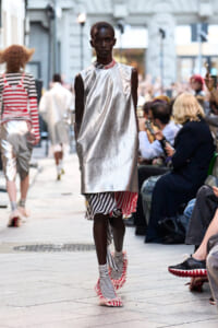 Dark-skinned model walks a street runway wearing a metallic silver sleeveless tunic over a striped layered skirt and fringed striped sandals, with spectators along the sides.