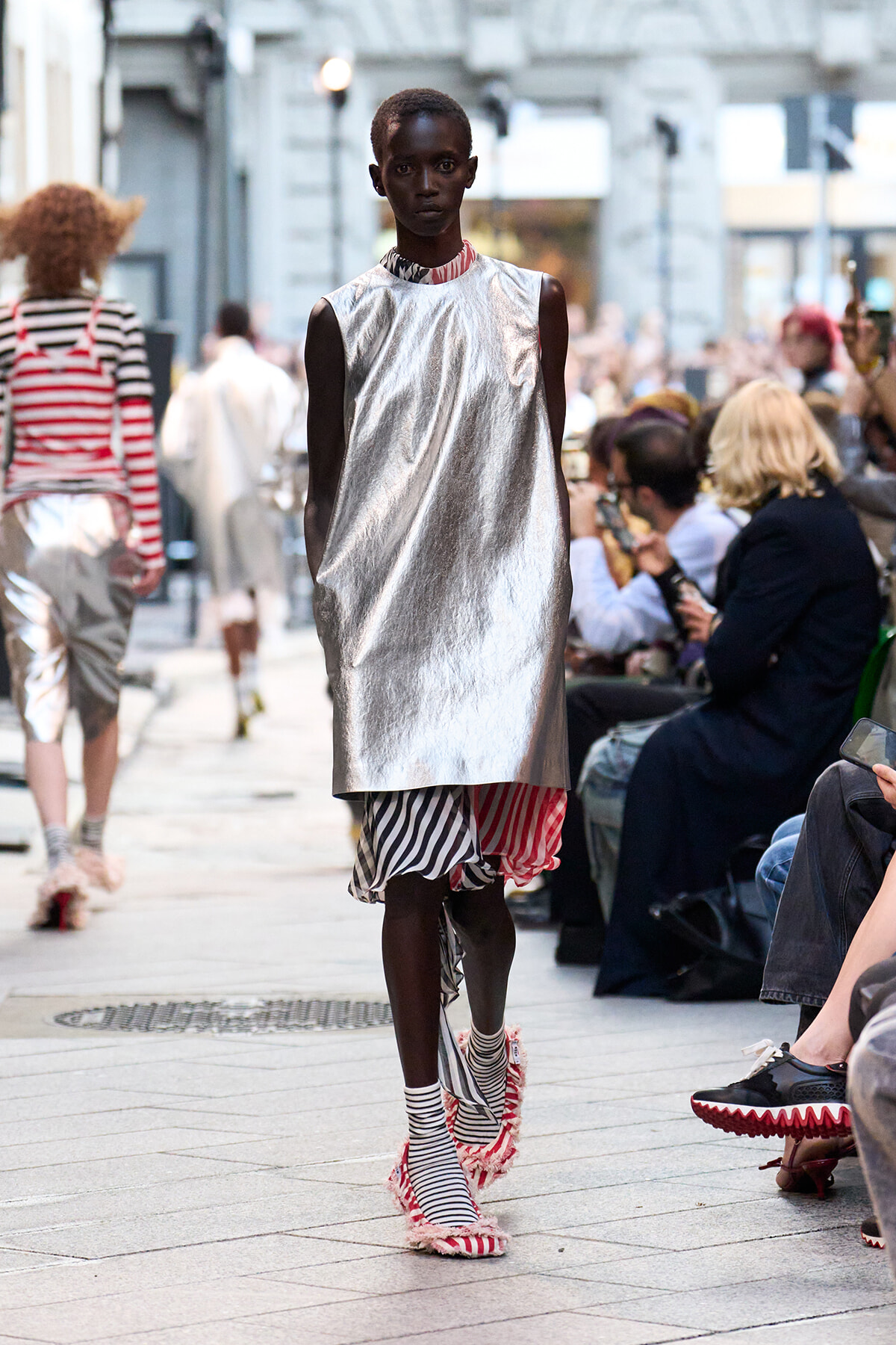 Dark-skinned model walks a street runway wearing a metallic silver sleeveless tunic over a striped layered skirt and fringed striped sandals, with spectators along the sides.