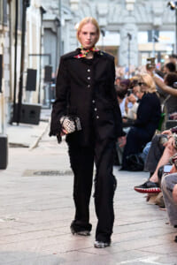 Model walking a street-style runway in a black fringed suit, accessorized with a colorful scarf and a polka-dot clutch, as spectators watch nearby.