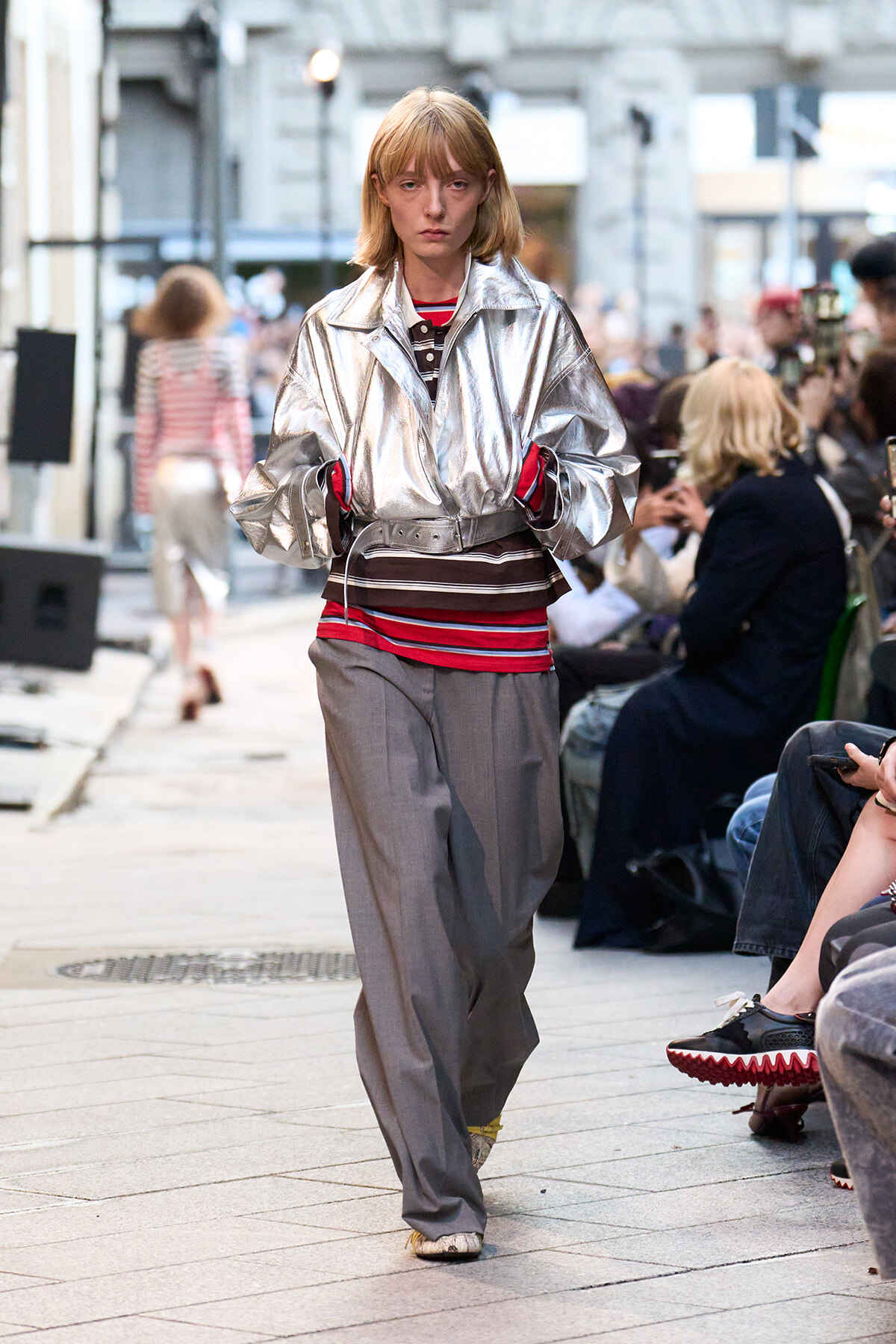Model walks a city street runway in a reflective silver oversized jacket, striped top, and wide gray trousers.