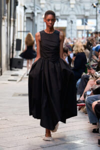 Model walks a street-style runway in a black sleeveless dress with a voluminous skirt, audience watching along the sides.
