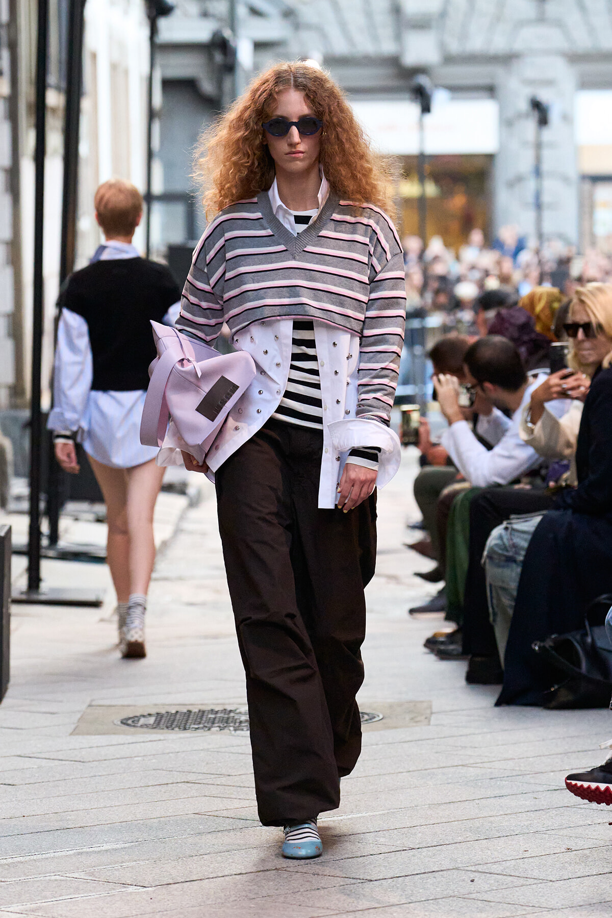 Fashion model with curly red hair wearing sunglasses walks a street-style runway, in a striped oversized sweater over a white shirt and dark wide-leg pants, carrying a pale pink bag.