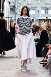 Model walking a street-style fashion show in a loose black-and-white striped top and a voluminous white skirt, with striped socks visible beneath the hem.