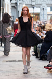 Model walking a street fashion show in a sleeveless black fringed dress with striped socks and metallic shoes on a crowded sidewalk runway.