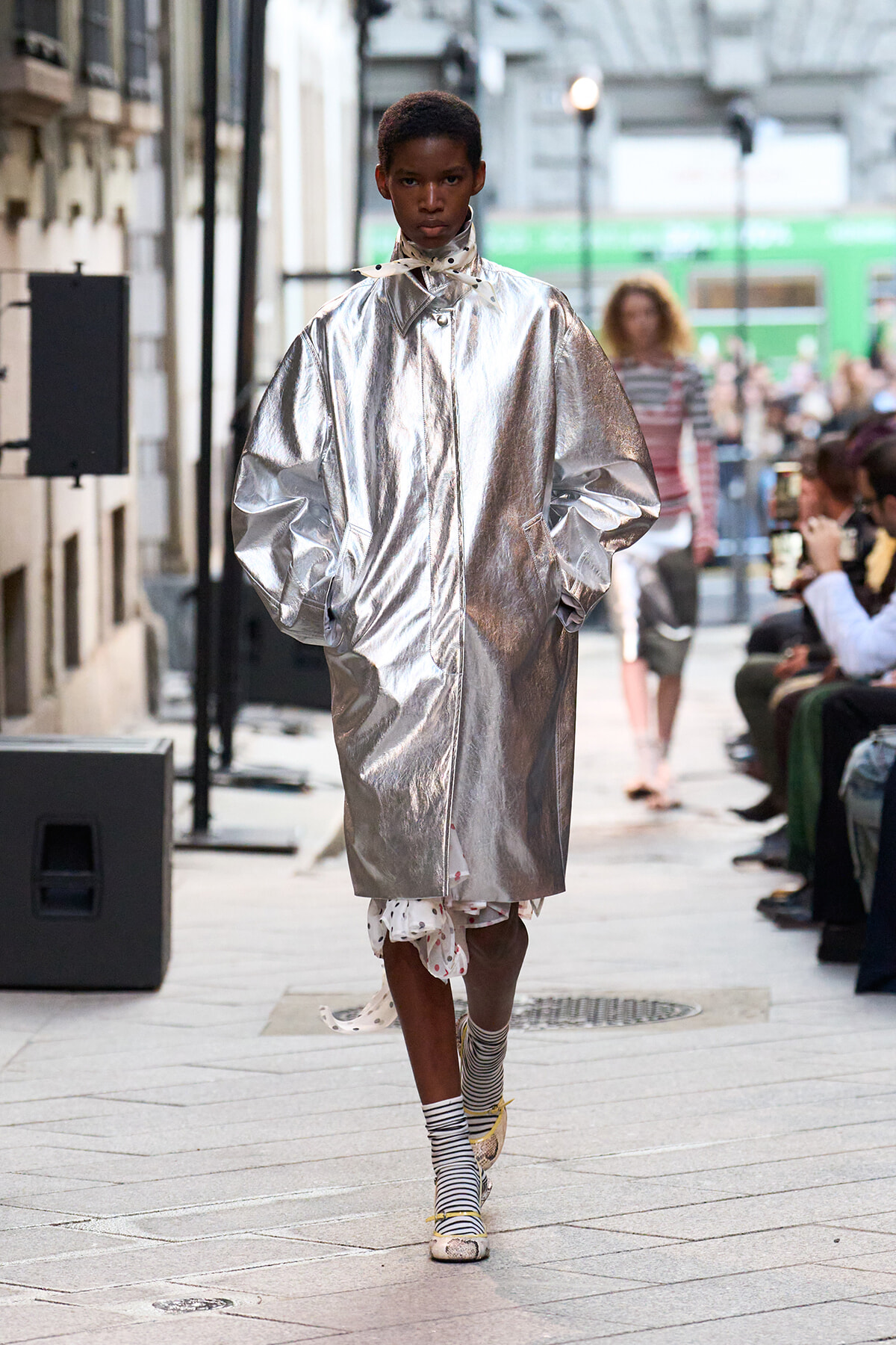 Model walks a street-style runway in a shiny oversized silver coat, hands in pockets, wearing striped socks and light-colored shoes. In the background, an audience watches.
