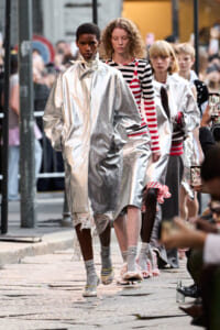 Models walk a street-fashion runway in metallic silver coats amid a crowd filming with smartphones.