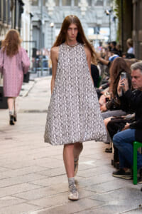 Model walks a snake-print A-line dress down a city street runway, audience seated on either side.