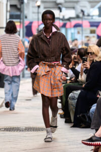 Model walking a street-style runway in a brown patterned jacket, pink shirt collar, orange plaid wrap skirt with a belt, wearing striped socks.