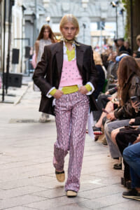 Model walks a street-style runway wearing a black blazer over a lime green shirt, pink beaded vest, and snakeskin-pattern pink pants with white shoes; audience seated on sides.
