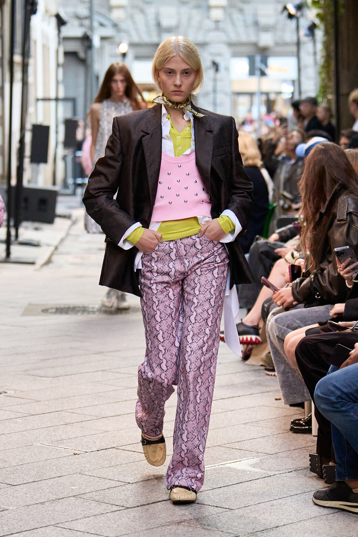 Model walks a street-style runway wearing a black blazer over a lime green shirt, pink beaded vest, and snakeskin-pattern pink pants with white shoes; audience seated on sides.