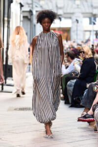 Model walks a street fashion show in a sleeveless black-and-white striped halter maxi dress, audience on both sides.